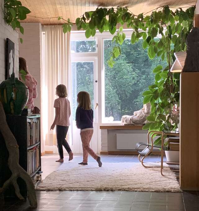 Kaksi lasta kävelee huoneessa, jossa iso ikkuna ja vehreä kasvi.