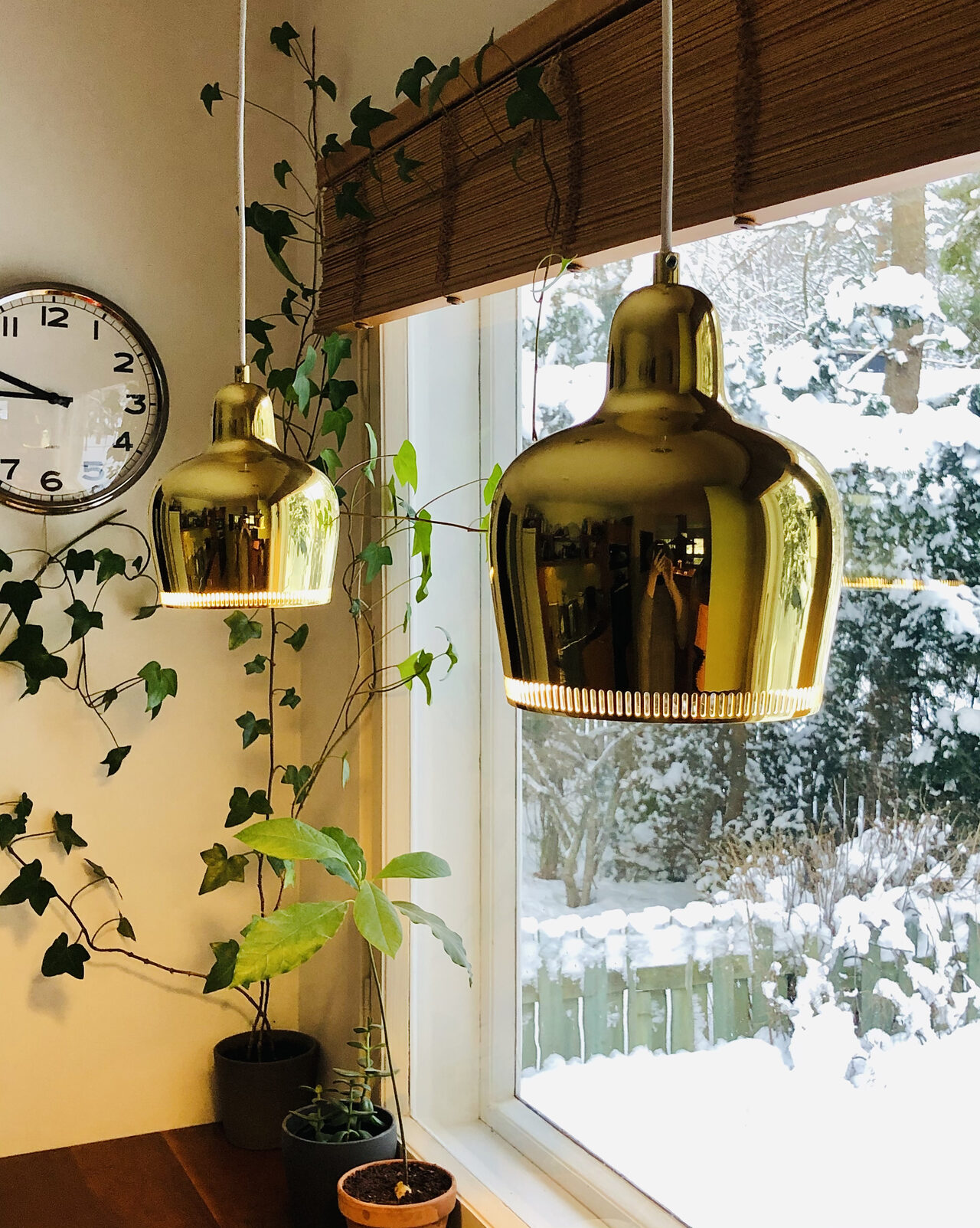 Kaksi kullanväristä valaisinta roikkuu katosta ikkunan edessä puisen työtason päällä. Taustalla köynnöskasvi ja seinäkello.