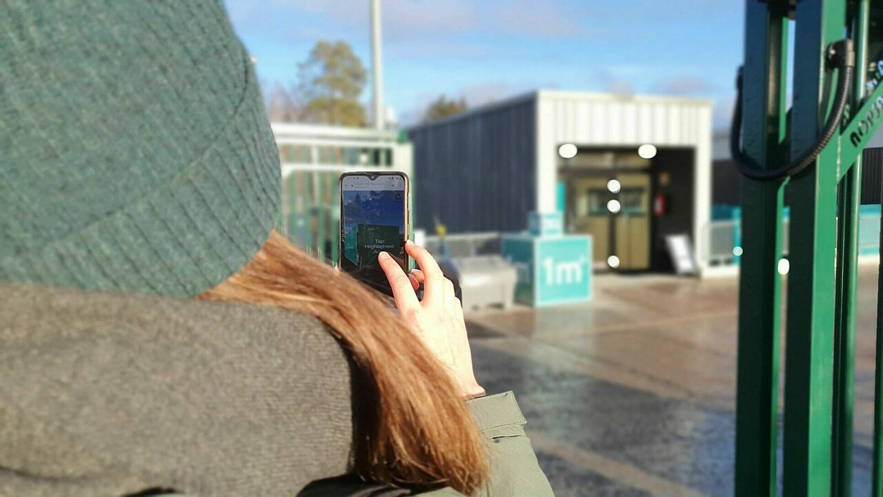 Pipopäinen henkilö katsoo älypuhelimen näytön läpi taaempana näkyvää laatikkomaista rakennusta.