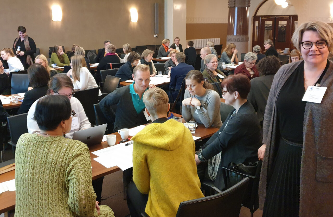 Apolityöpaja Sonckin salissa, jossa ihmiset ovat keräytyneet keskustelemaan pöytien ääreen. Kuvan etualalla on Leena Rossi.