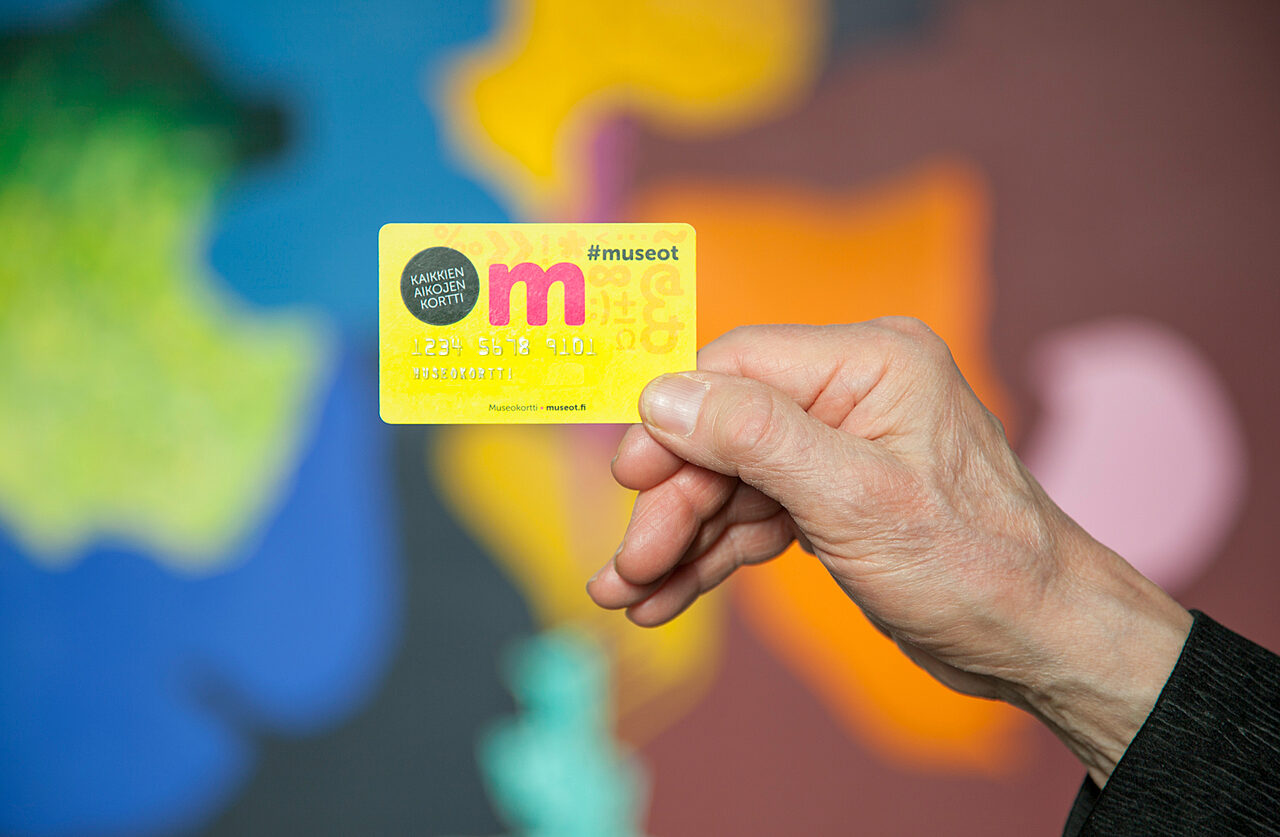 Käsi pitää keltapinkkiä museokorttia ilmassa. Taustalla abstrakteja ja värikkäitä kuvioita.