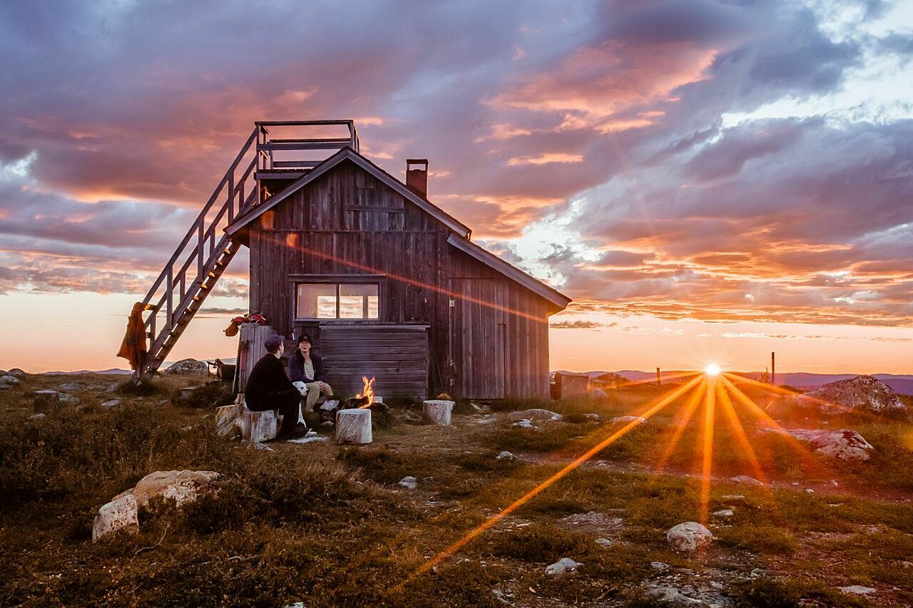 Kuva puurakennuksesta, jonka edustalla kaksi ihmistä ovat nuotiolla