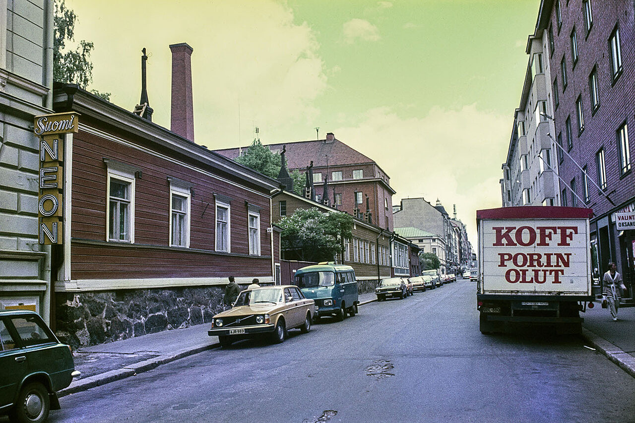 Vanha värikuva kadusta, vasemmalla matala puutalo, oikealla tiilikerrostalo, kadulla autoja ja Koffin kuorma.auto