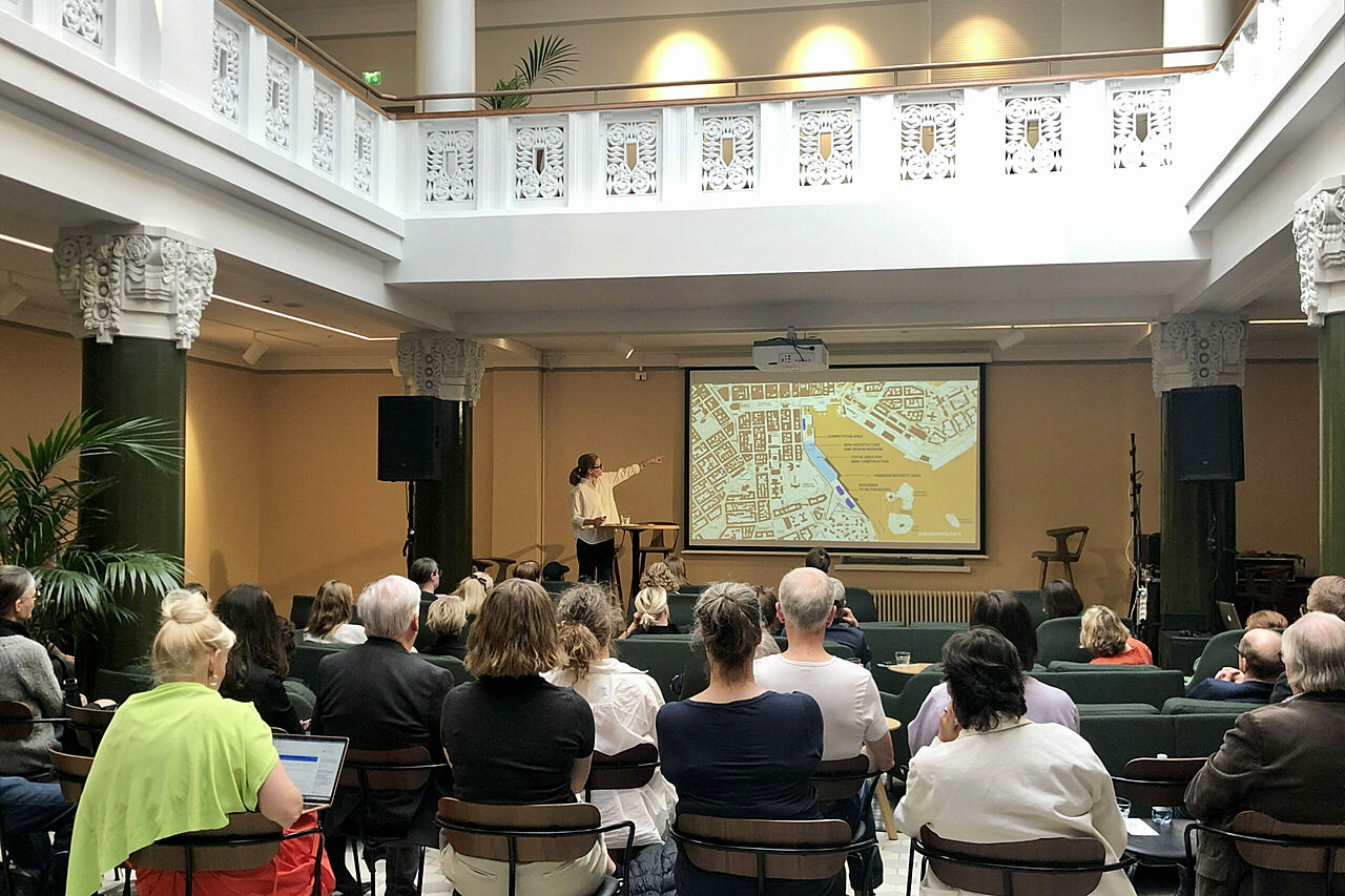 Korkea tila, jossa pilarit ja koristeellinen parvikerros. Edessä nainen osoittaa valkokangasta, yleisö istuu.