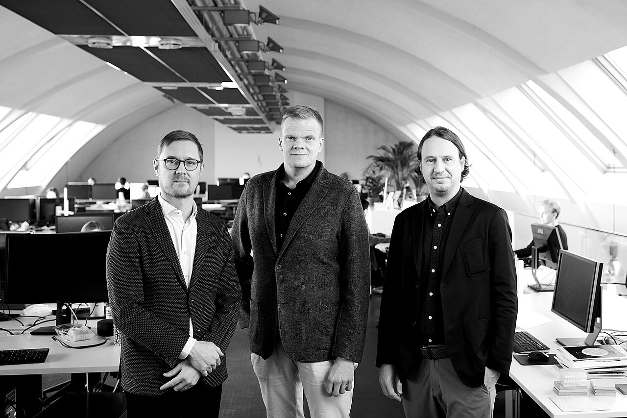 Kolme miestä katsoo kameraan toimistotilassa, jossa on kaareva katto.