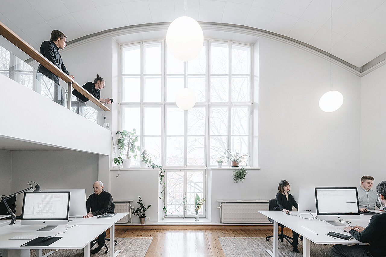 Sisäkuva korkeasta valkoisesta tilasta, taustalla kaari-ikkuna, vasemmalla parvi. Ihmisiä työskentelee työpöytien ja tietokoneiden ääressä.