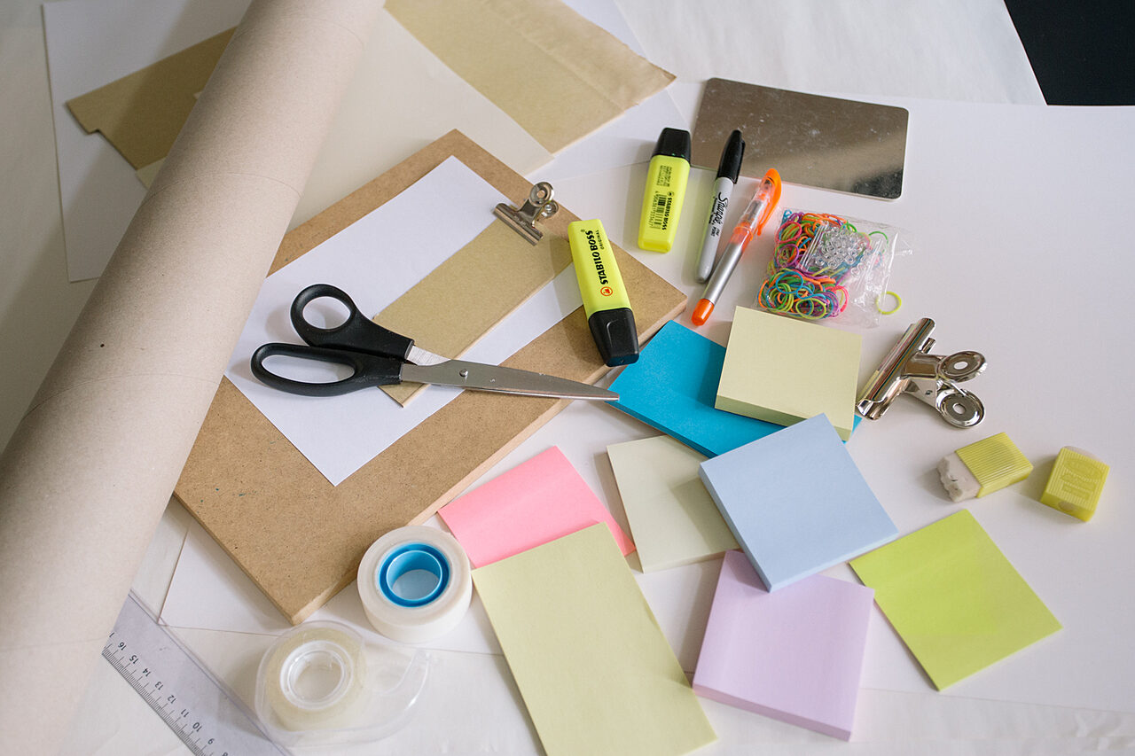 Kuva työpöydästä, jonka päällä on pinossa post-it lappuja, sakset, yliviivaustusseja, teippejä ynnä muuta.