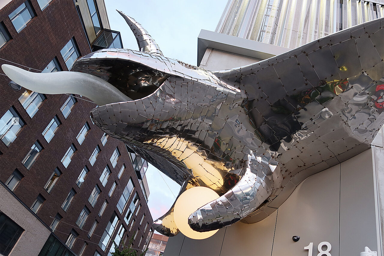 Talon ulkonurkassa liskon näköinen metalliveistos, jolla pitkä kieli ja pyöreä valaisin käpälien välissä.