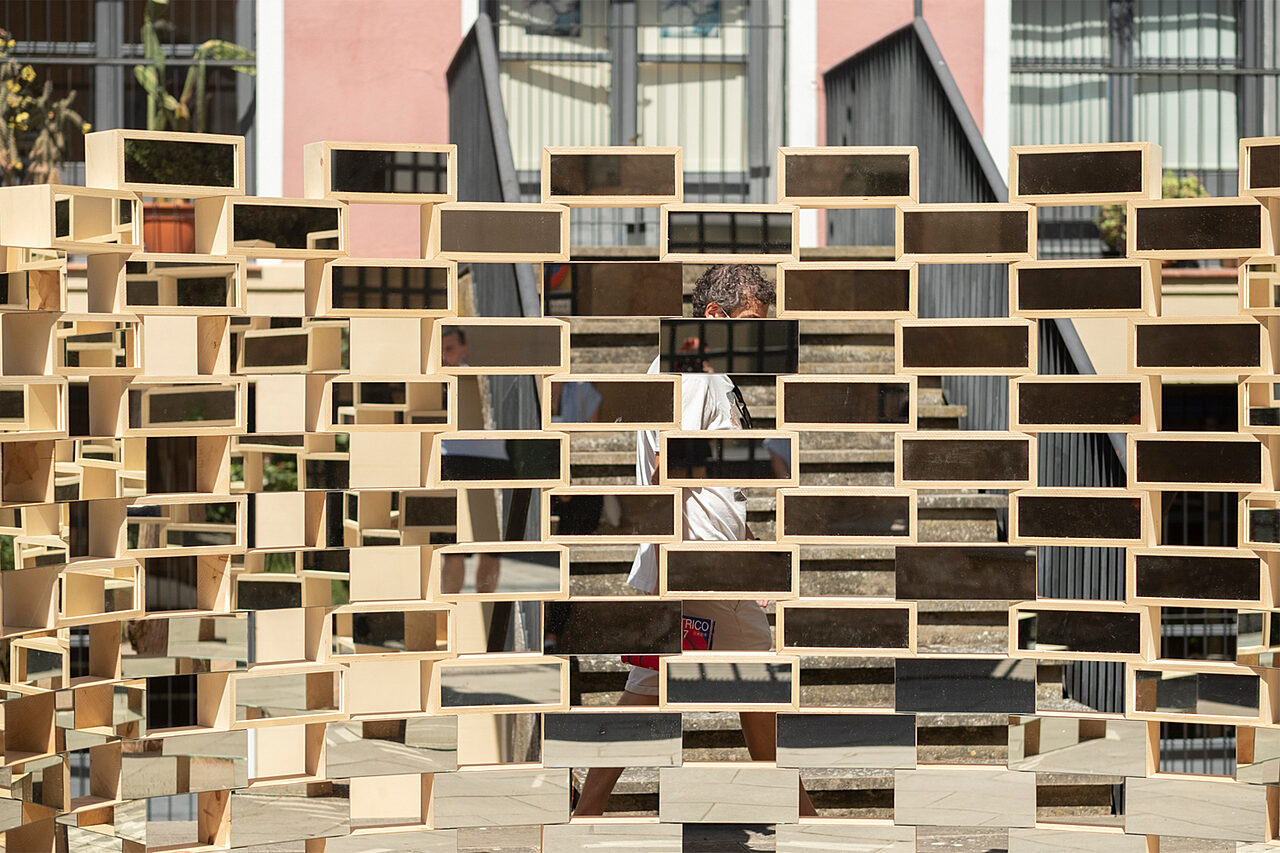 Puisia vähän tiiltä suurempia laatikoita on aseteltu limittäin toistensa päälle. Laatikoiden katsojan puoleinen sivu on peiliä. Laatikoiden välisistä aukoista näkyy kävelevä mies.