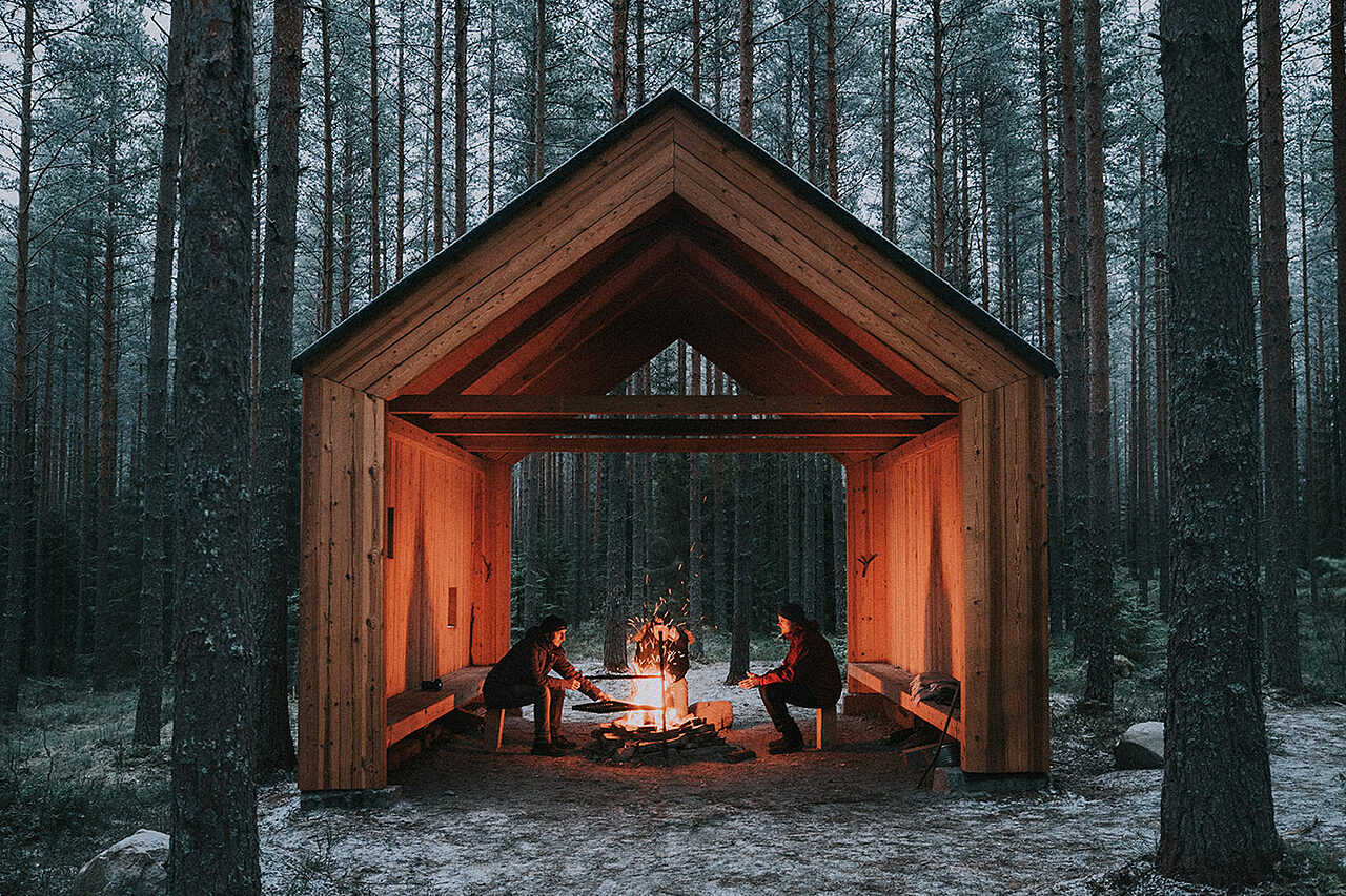 Puinen nuotiokatos metsän keskellä, katoksessa palaa nuotio, jonka äärellä istuu kaksi ihmistä.