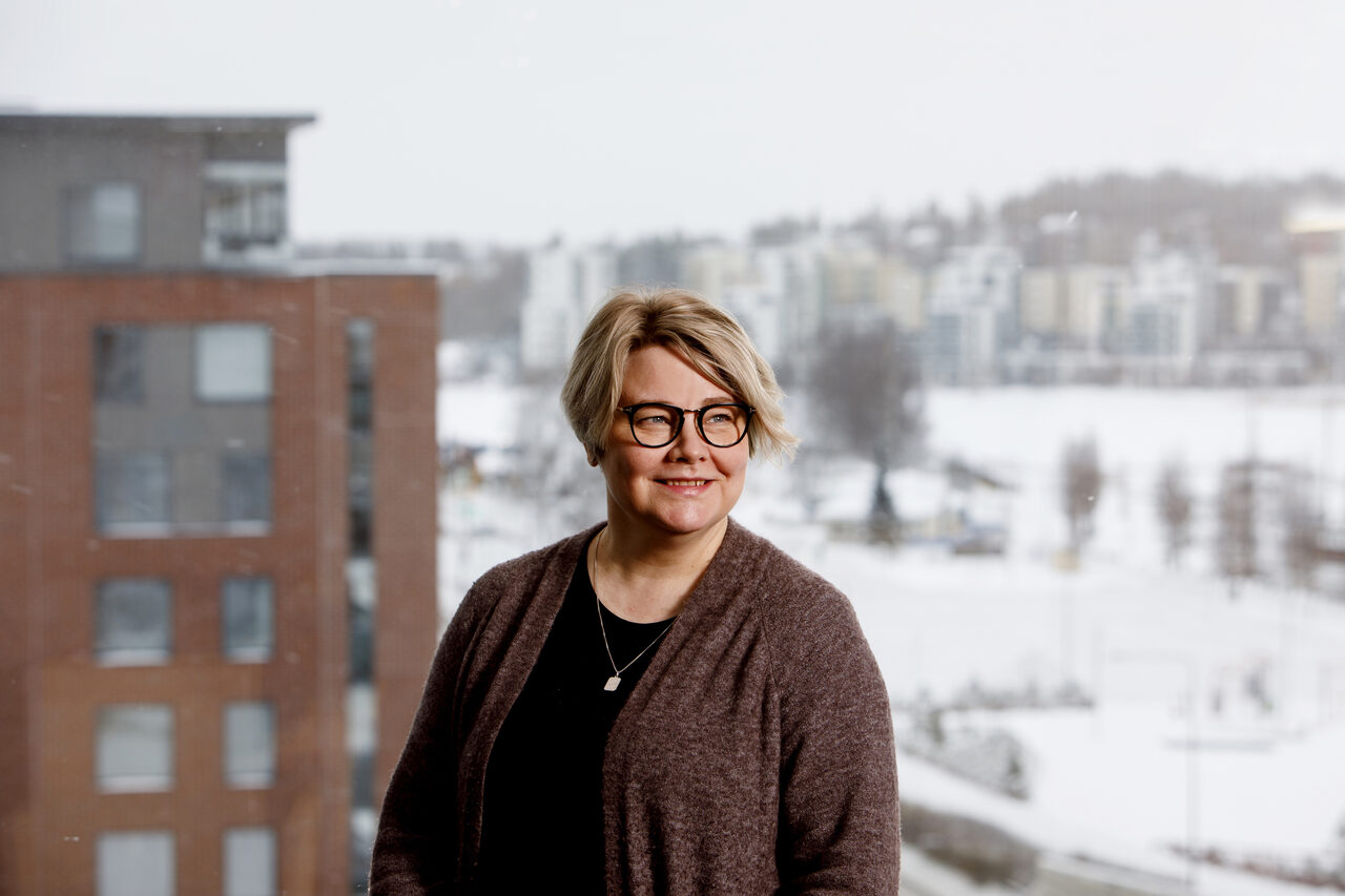 Nainen katsoo sivulle. Kuvan taka-alalla on talvinen maisema sekä punertava kerrostalo