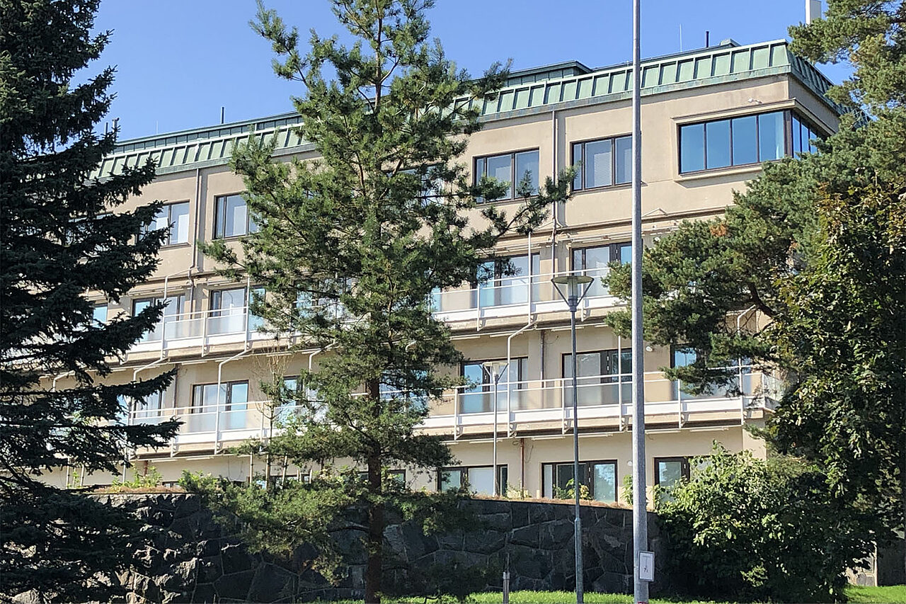 Nelikerroksinen rakennus kuvattuna puistosta päin. Vaaleanruskean rakennuksen kahta keskimmäistä kerrosta kiertävät parvekkeet.