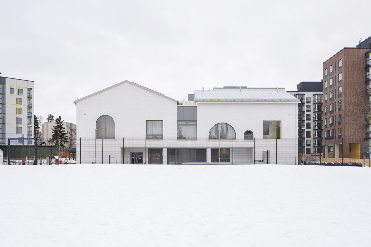 Martinlaakson päiväkoti: valkoinen kaksikerroksinen rakennus, joka näyttää koostuvan kahdesta osiosta, joista toisen harjakatto osoittaa katsojaan päin ja toisen katsojasta oikealle. Rakennuksen yläkerran neljästä ikkunasta kaksi ovat päältä kaarevia ja kaksi neliskulmaisia.