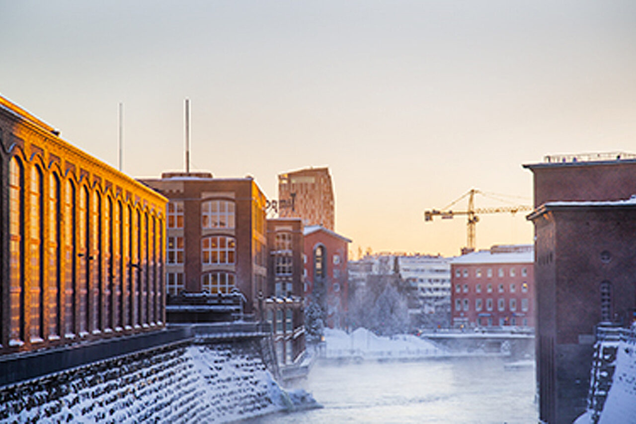 Punatiilisiä rakennuksia kosken molemmin puolin. Kosken rannoilla on lunta. Taustalla kohoaa huomattavan korkea kerrostalo sekä nostokurki.
