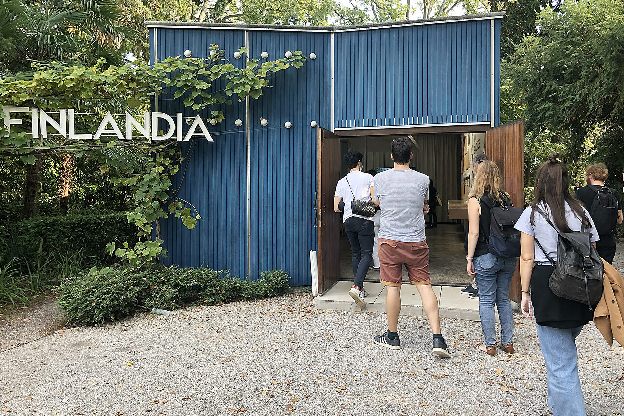 Kesäisiä ihmisiä jonottaa sisään pieneen siniseen puurakennukseen, jonka kyljessä roikkuu teksti Finlandia.