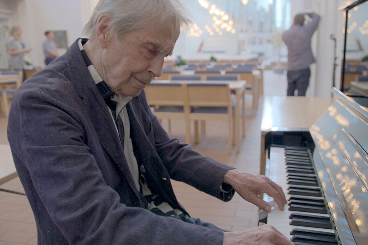 Harmaahiuksinen mies soittaa pianoa valoisassa tilassa.