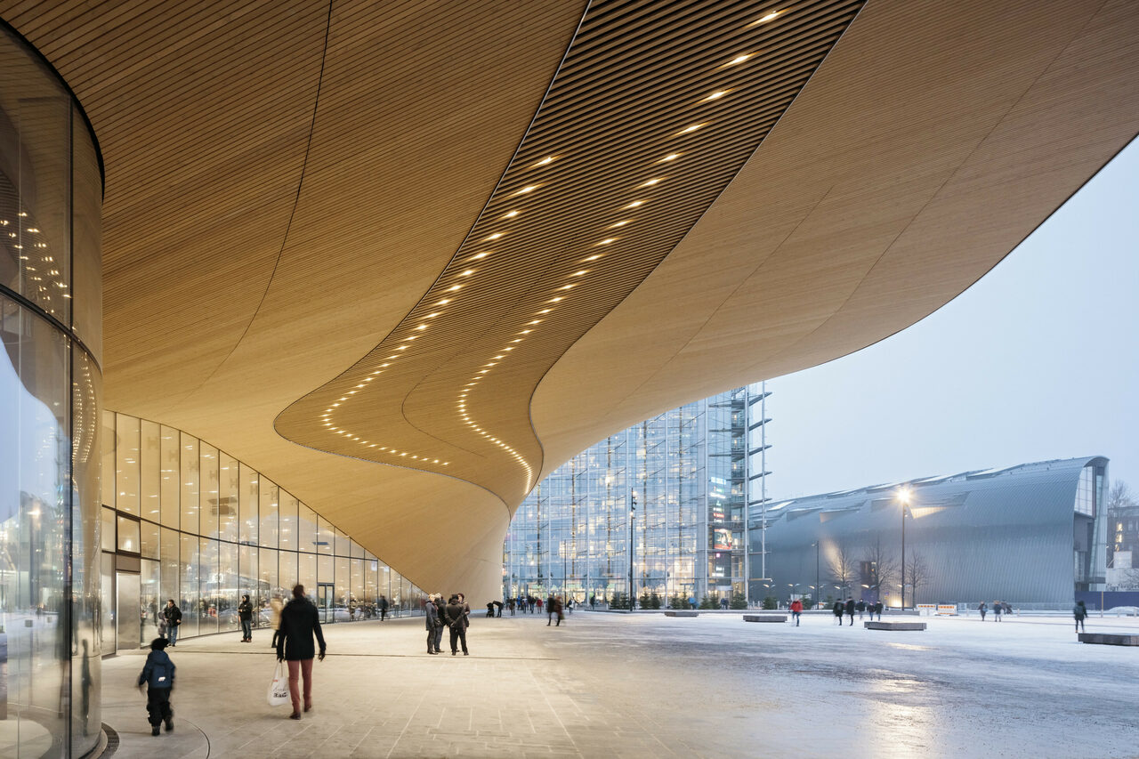 A curved wooden canopy covering the entrance to a big building on a square, some people coming and going