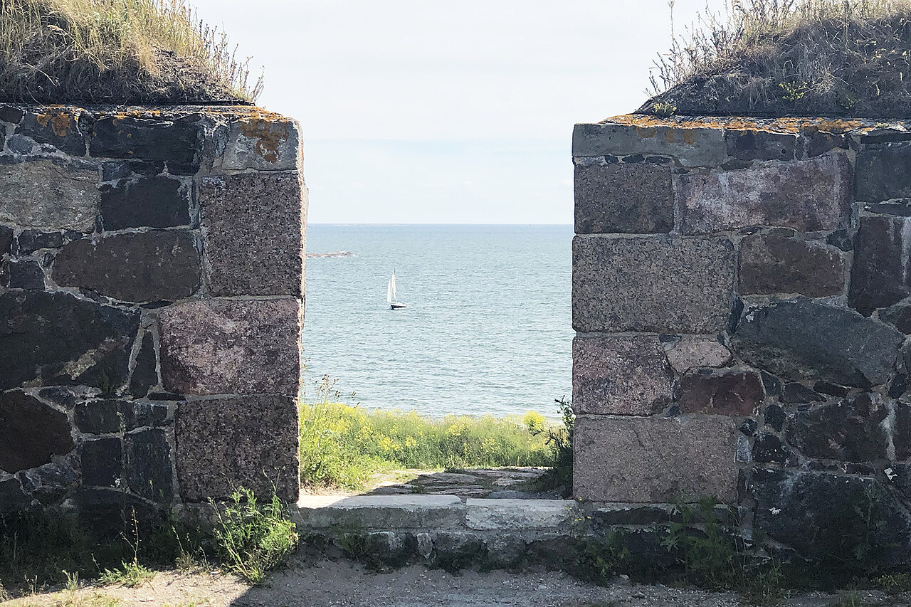 Turvekattoisten graniittimuurien välistä näkyy aurinkoinen ulappa ja siellä yksi purjevene
