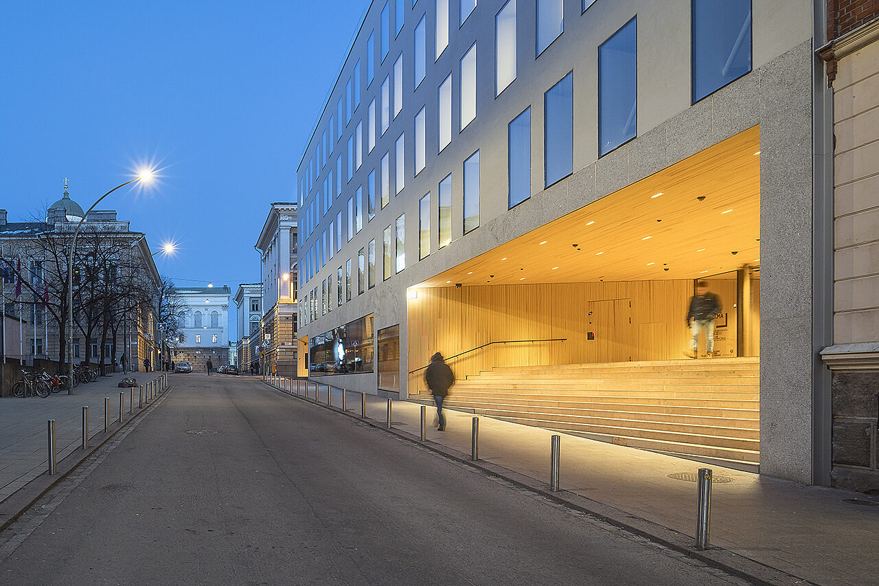 Iltakuva, nouseva katu, oikealla valoisa puupäällysteinen sisäänkäyntisyvennys valkoisessa talossa, jossa paljon ikkunoita.