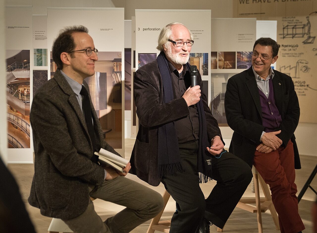 Three men are sitting on chairs in front of the exhibition.