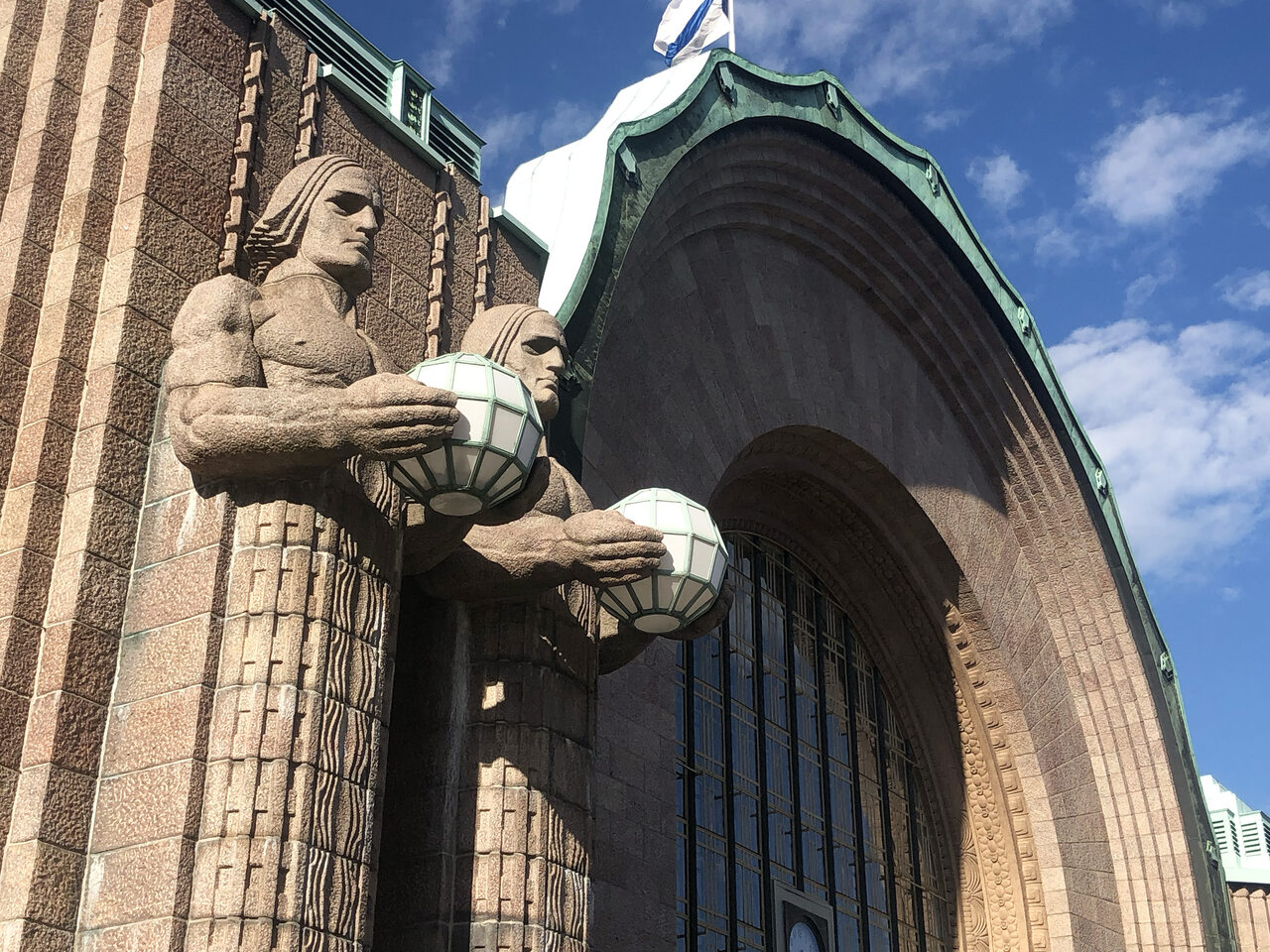 Katse ylöspäin kohti graniittisen rakennuksen seinästä ulos työntyviä patsaita miehistä, jotka kannattelevat lasipalloja.