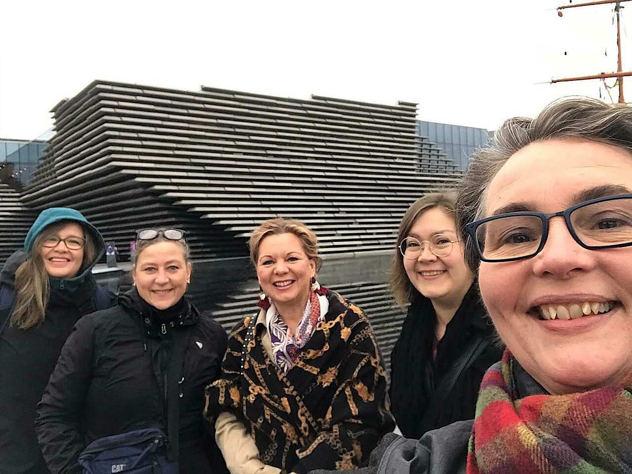 Viisi naista poseeraa hymyillen kameralle. Taustalla näkyy V&A Dundee -deisgnmuseon julkisivua.