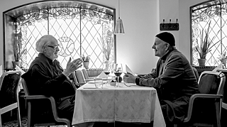 Two men sit facing each other around a table, engaged in conversation.