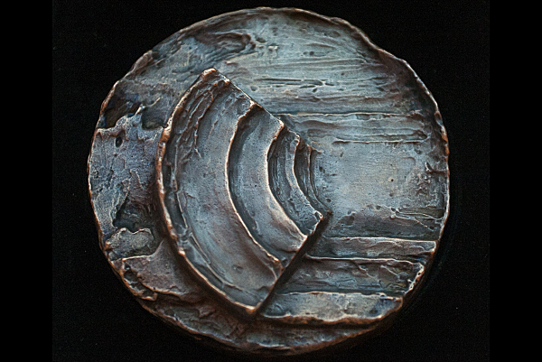 Pyöreä, sinertäväksi patinoitunut kuparinen mitali, jonka keskellä on amfiteatteria muistuttava muoto.