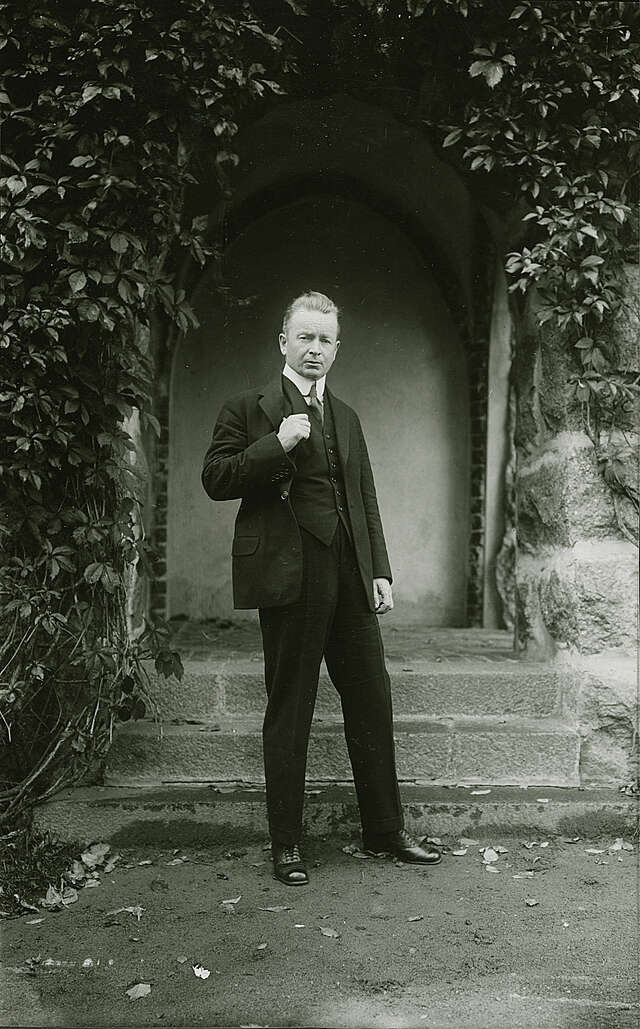 Vanha mustavalkoinen valokuva siistiin pukuun pukeutuneesta miehestä, joka seisoo köynnöskasvien ympäröimän sisäänkäynnin edessä.