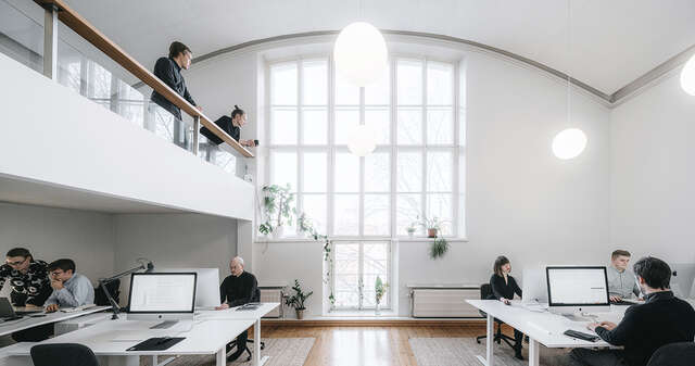 A white atelier space with a high curved ceiling. People at their desks and on the mezzanine.