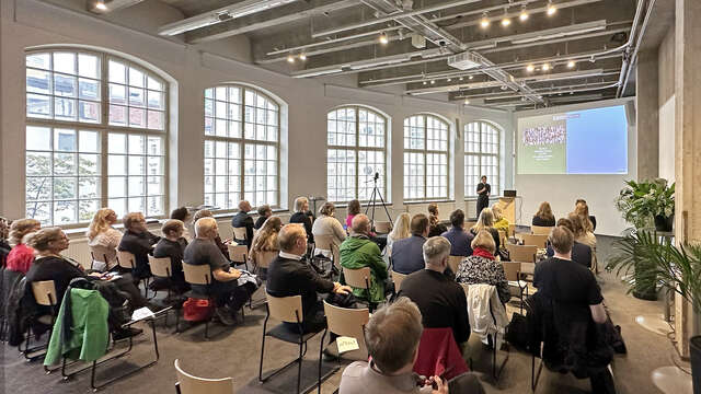 Huone täynnä yleisöä istumassa ja kuuntelemassa esitelmää.