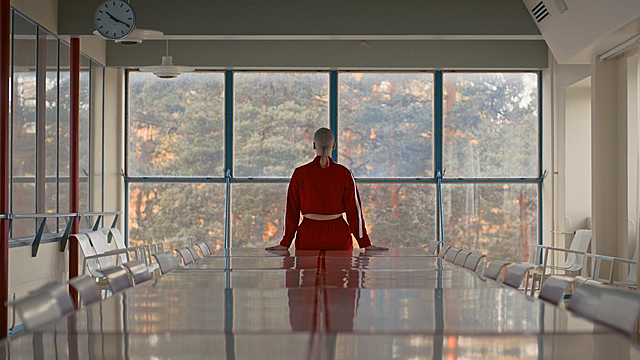 Naisoletettu nojaa suureen kiiltävään pöytään selin kameraan, takaseinä ikkunaa