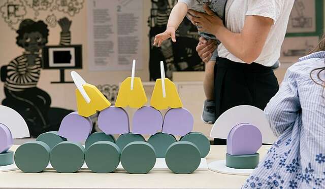 Vihreitä, violetteja ja keltaisia kappaleita pöydällä, taustalla lapsi aikuisen sylissä