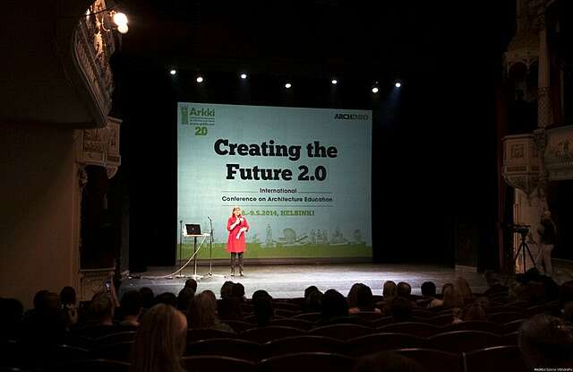 Nainen puhuu lavalla suuren yleisön edessä pimennetyssä huoneessa.