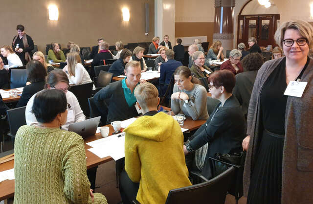 Apolityöpaja Sonckin salissa, jossa ihmiset ovat keräytyneet keskustelemaan pöytien ääreen. Kuvan etualalla on Leena Rossi.