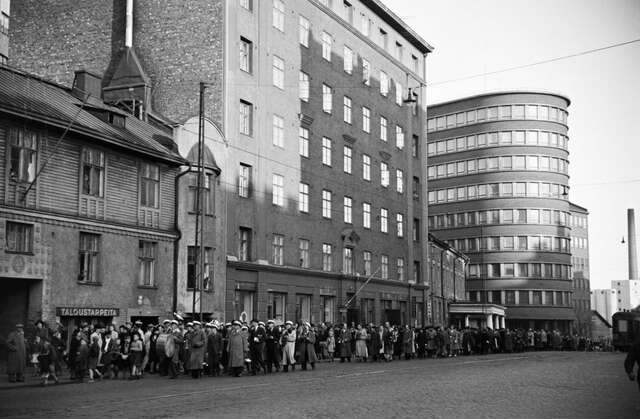 BW photo, long procession of people walking in the street, red brick building with a rounded end in the background