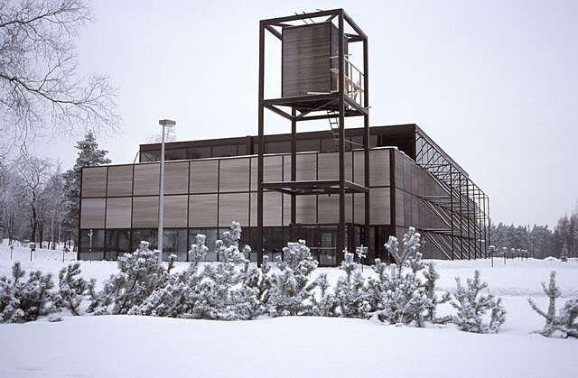 Talvinen maisemakuva Kouvolan keskuskirkosta, joka on moderni, vaalea kirkko. Etualalla näkyy lumisia pensaita ja kirkon kulmasta nouseva korkea, metallirakenteinen kellotorni harmaata talvitaivasta vasten.