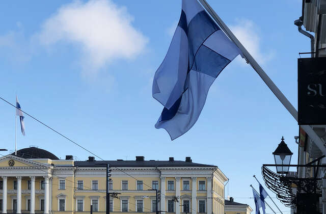 Suomen lippu etualalla, taustalla keltainen talo.