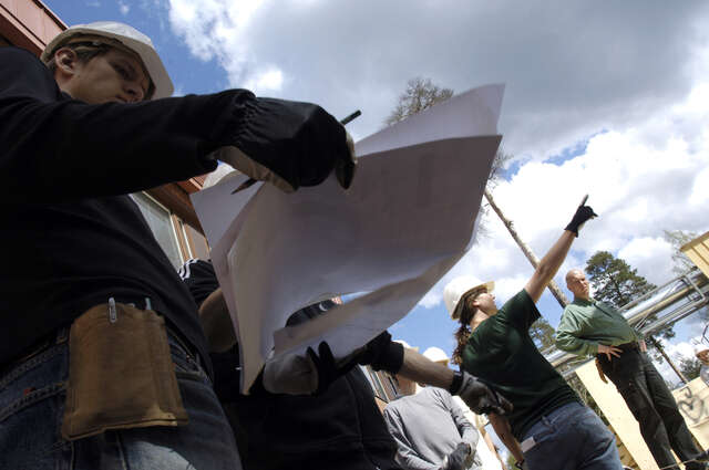 Näkymä alaviistosta ihmisiin, joista osa katsoo kädessään olevia papereita. Ihmisillä on päässään kypärät.