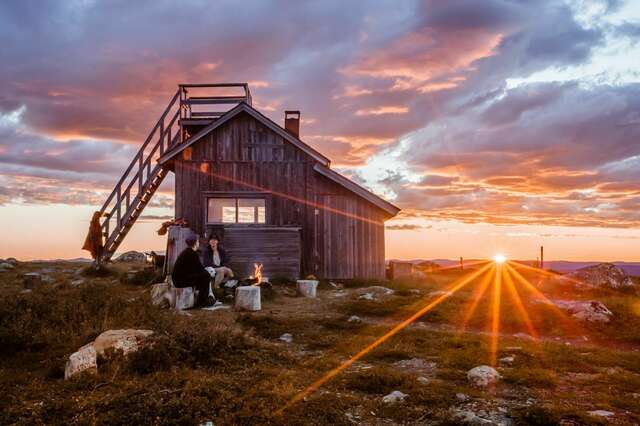 Picture of a wooden building with two people around a campfire in front of it