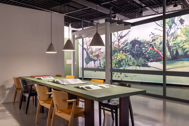 Kuva näyttelytilasta, jossa on ruokapöytä ja ikkuna kuvitteelliseen, maalauksella toteutettuun puutarhaan