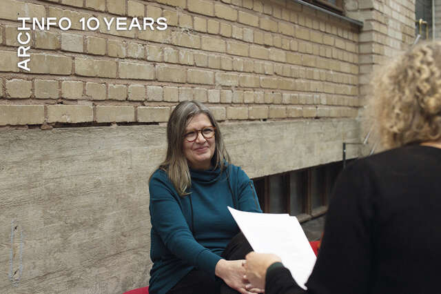 Nainen turkoosinsinisessä paidassa istuu vastapäätä vaaleaa kiharatukkaista naista, taustalla keltatiiliseinä