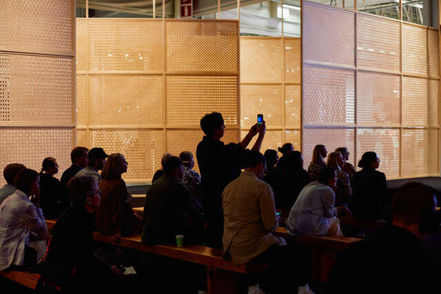 The crowd is turned toward a stage. Wooden room dividers are in the background.