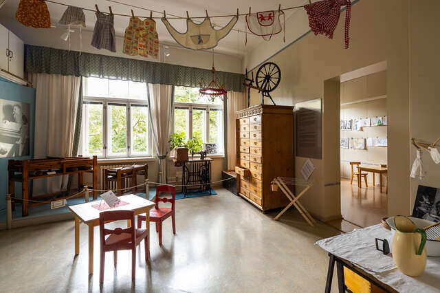 Sisäkuva huoneesta vanhassa talossa, jossa suuret kolmijaolliset ikkunat. Punaiset puutuolit ja pöytä etualalla, ylhäällä roikkuu narulla vaatteita.