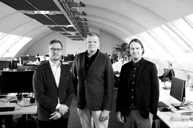 Three men look at the camera in an office space with a vaulted ceiling.