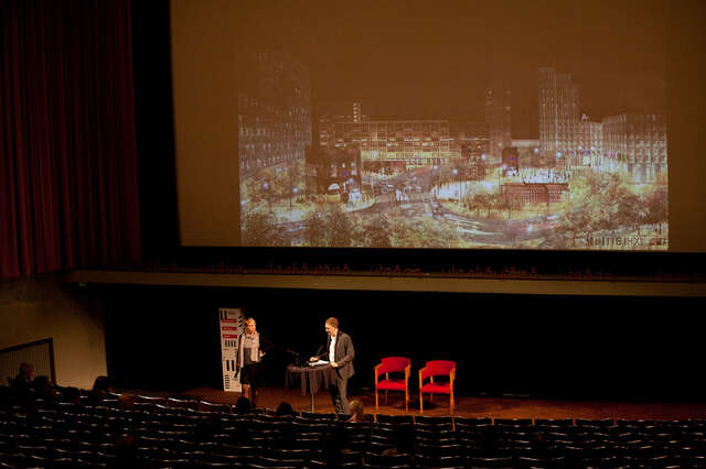 Mies puhuu suuren yleisön edessä, näytöllä näkyy kaupunkimaisema.