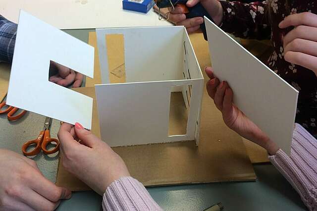 Pöydällä on pienoismalli, jota kootaan. Kuvassa näkyy käsiä, mutta ei itse ihmisiä.