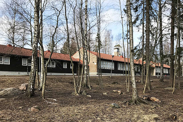 Ohuiden koivunrunkojen takana pitkä kolmiosainen koulun julkisivu. Punainen tiilikatto, tummat lautavuoratut seinät, valkoiset ikkunankehykset.