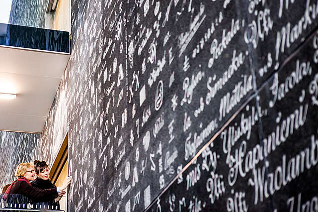 Kaksi naista ihoilee Kansallisarkiston Hämeenlinan toimipaikan seinää, jossa on kirjoitusta eri kielillä ja merkeillä.