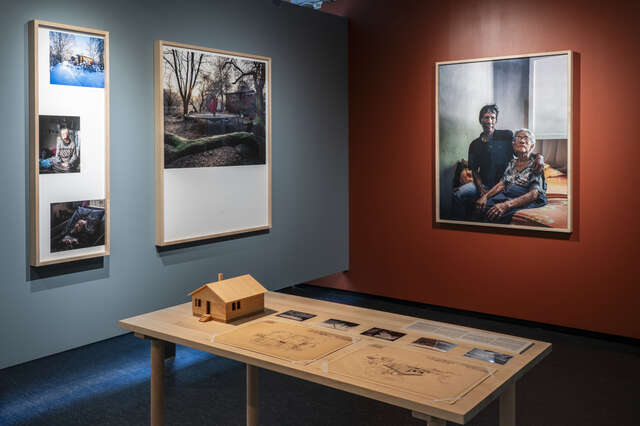 Näyttelytila, jossa esillä valokuvia seinällä ja pöytä, jonka päällä piirustuksia ja pienoismalli puutalosta.
